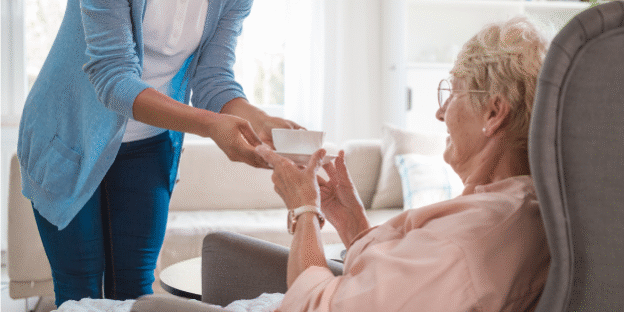 A caregiver hands a cup to an elderly woman sitting in a care home, representing the support and financial considerations involved in care home fee assessments in England.