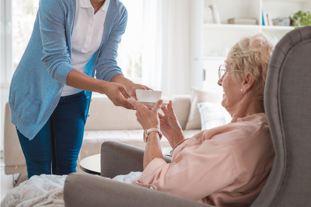 A caregiver hands a cup to an elderly woman sitting in a care home, representing the support and financial considerations involved in care home fee assessments in England.
