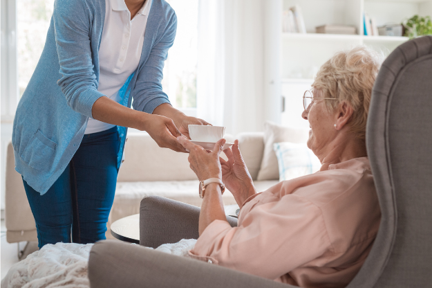 PHH Solicitors Fylde Coast A caregiver hands a cup to an elderly woman sitting in a care home, representing the support and financial considerations involved in care home fee assessments in England.
