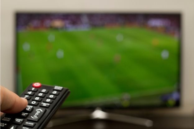 Hand holding a TV remote while watching a blurred football match, illustrating rising domestic abuse reports during World Cup events and the need for legal support in Lancashire.