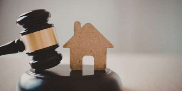 Wooden auction gavel next to a small house-shaped cutout on a sound block, representing buying a property at auction.
