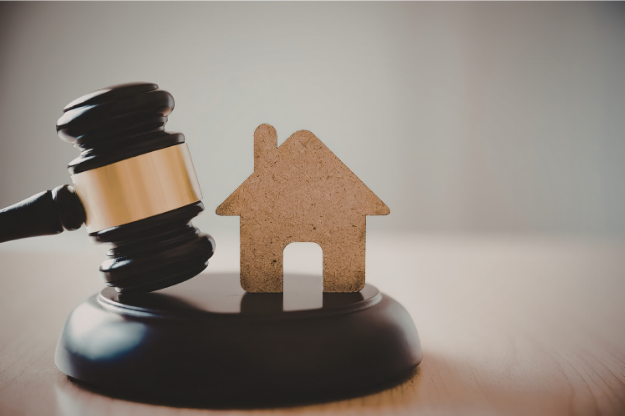Wooden auction gavel next to a small house-shaped cutout on a sound block, representing buying a property at auction.